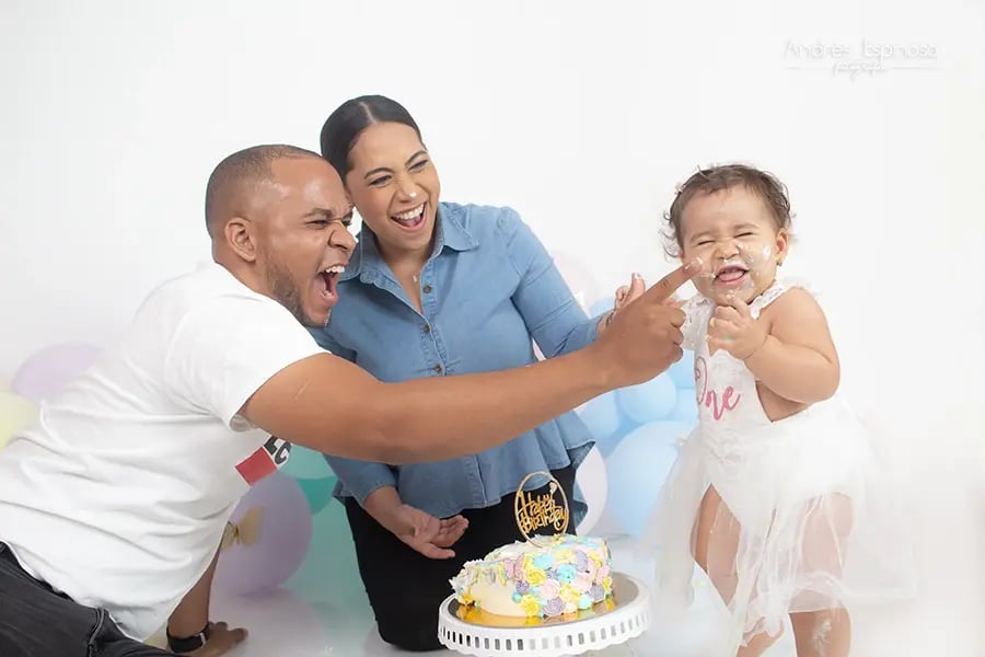 Familia celebrando el primer cumpleaños del bebé en una sesión smash cake. Padre y madre sonríen mientras el bebé se ensucia con el pastel.